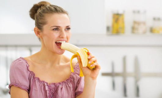 A woman wearing pink eats a banana in a bright room.