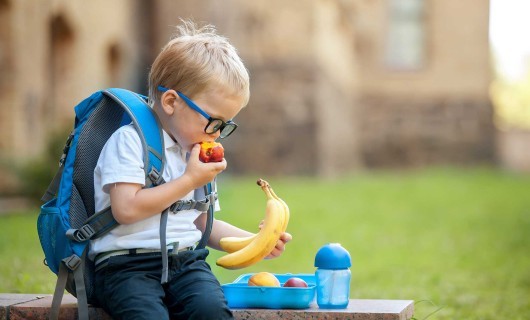 A young boy with glasses and a blue backpack is sat holding bananas and eating a nectarine.