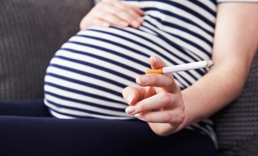 A heavily pregnant woman is sat holding her bump, with a cigarette in her hand.
