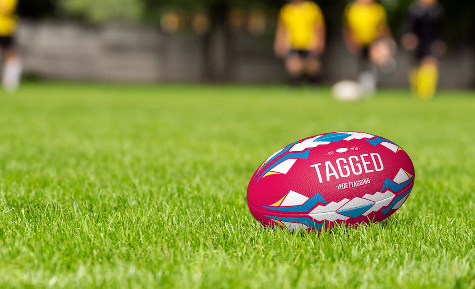 A pink rugby ball with Tagged branding on, in the grass.